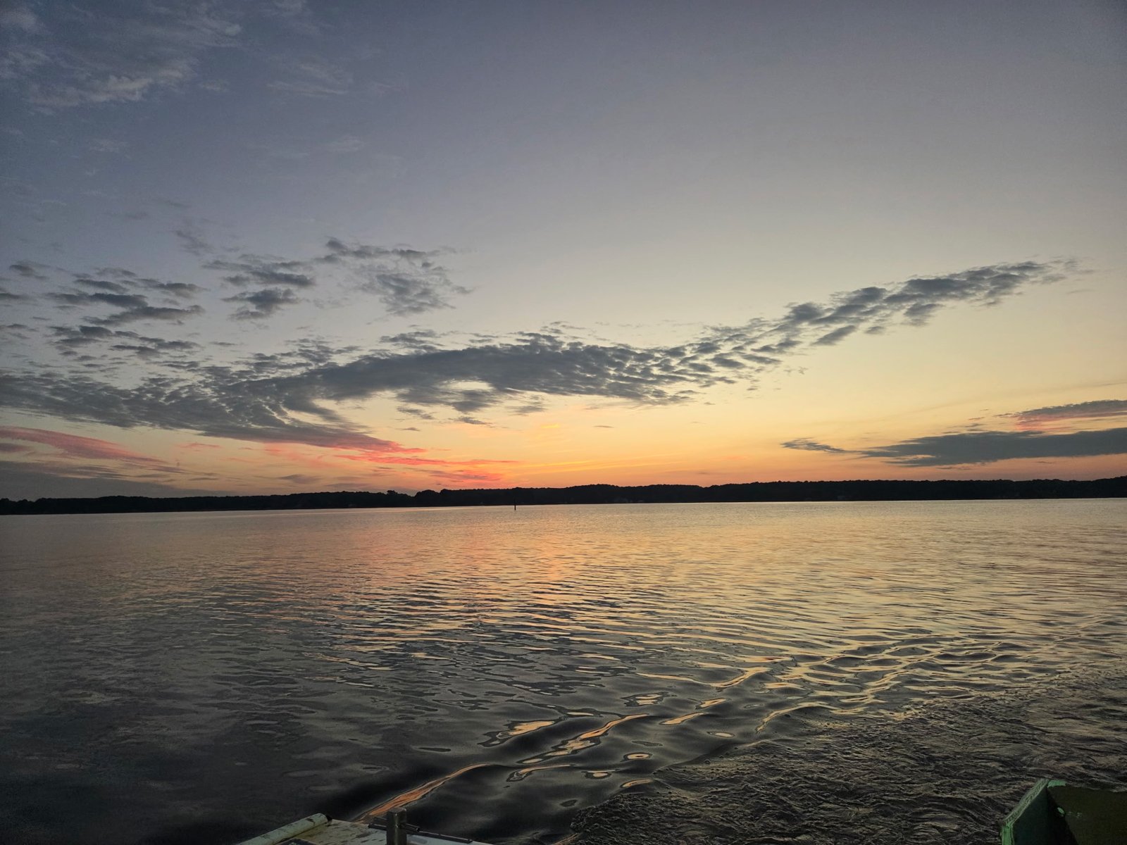 Dawn over open water, a low tree line on the far shore, orange and pink at the horizon, ripples in the foreground, the corner of a boat at the bottom