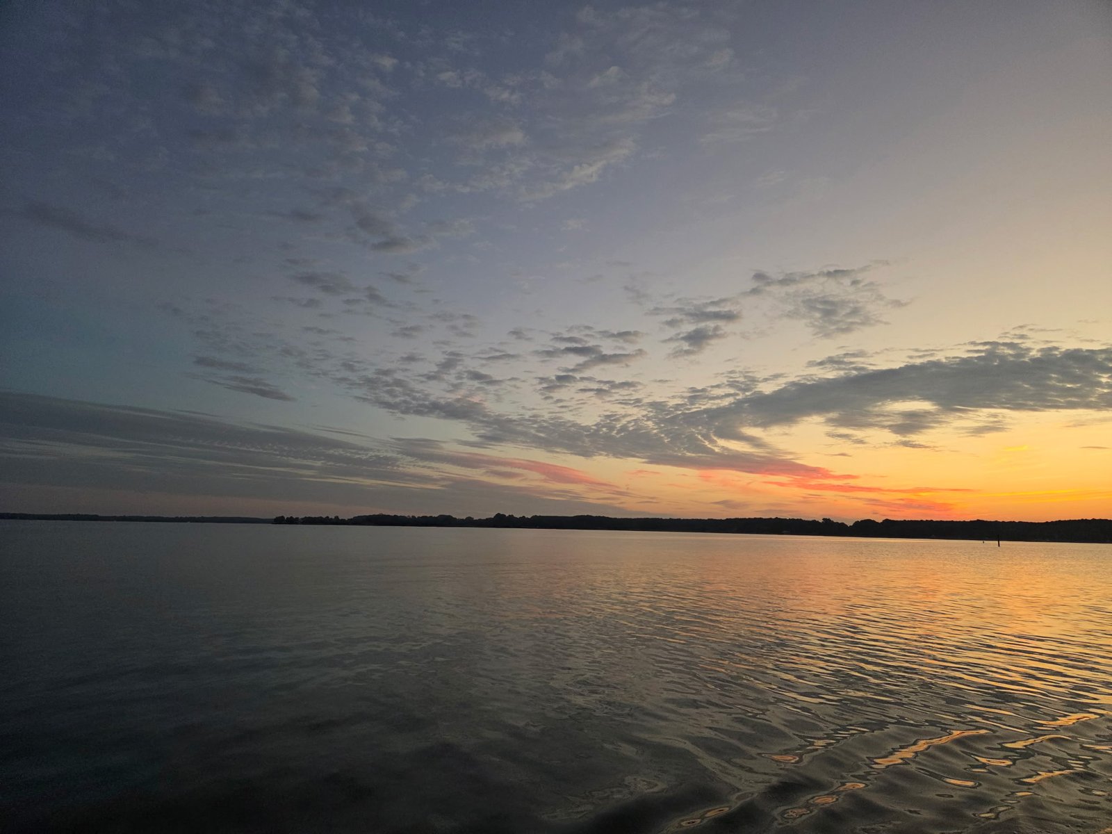 Calm water at dawn under a sky of small clouds, a pale blue-gray shading to orange at the horizon, a distant shoreline