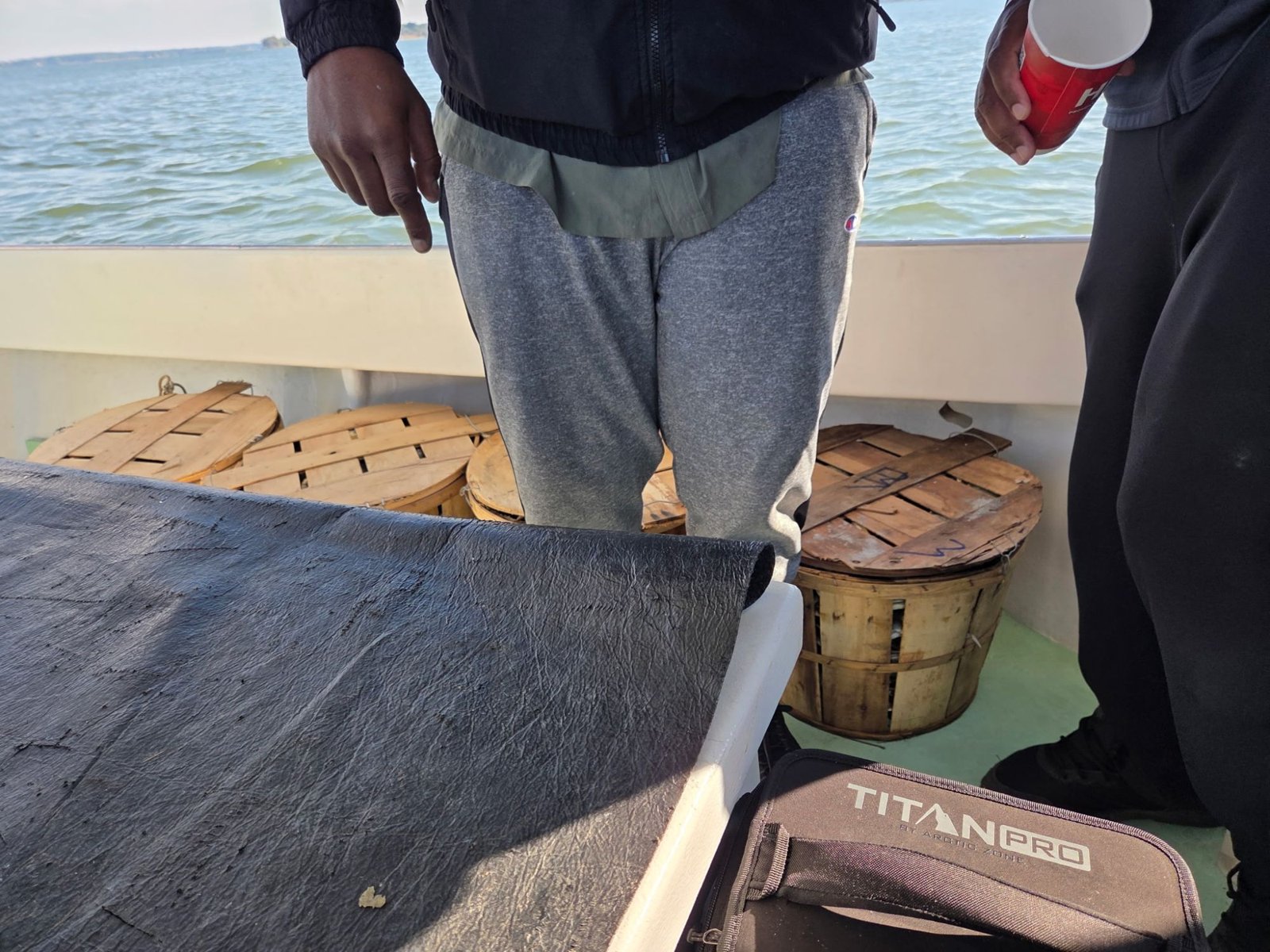 Wooden bushel baskets with lids on the deck of a boat, open water behind, two people standing partly out of frame