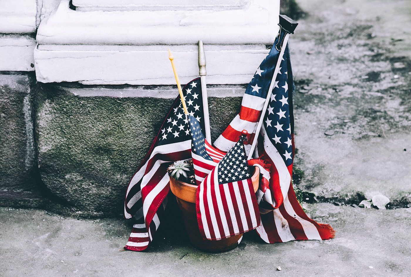 Several small, worn American flags leaning in a terracotta flowerpot at the base of a weathered stone wall