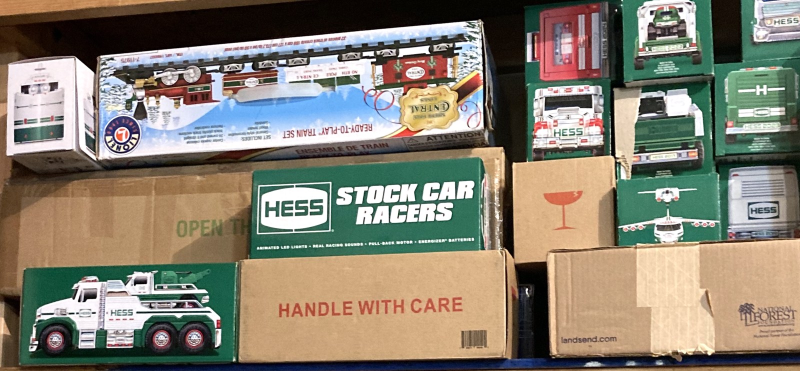 A shelf of Hess toy trucks in their green and white boxes, with a Hess tow truck box at lower left and a Hess stock car racers box in the middle