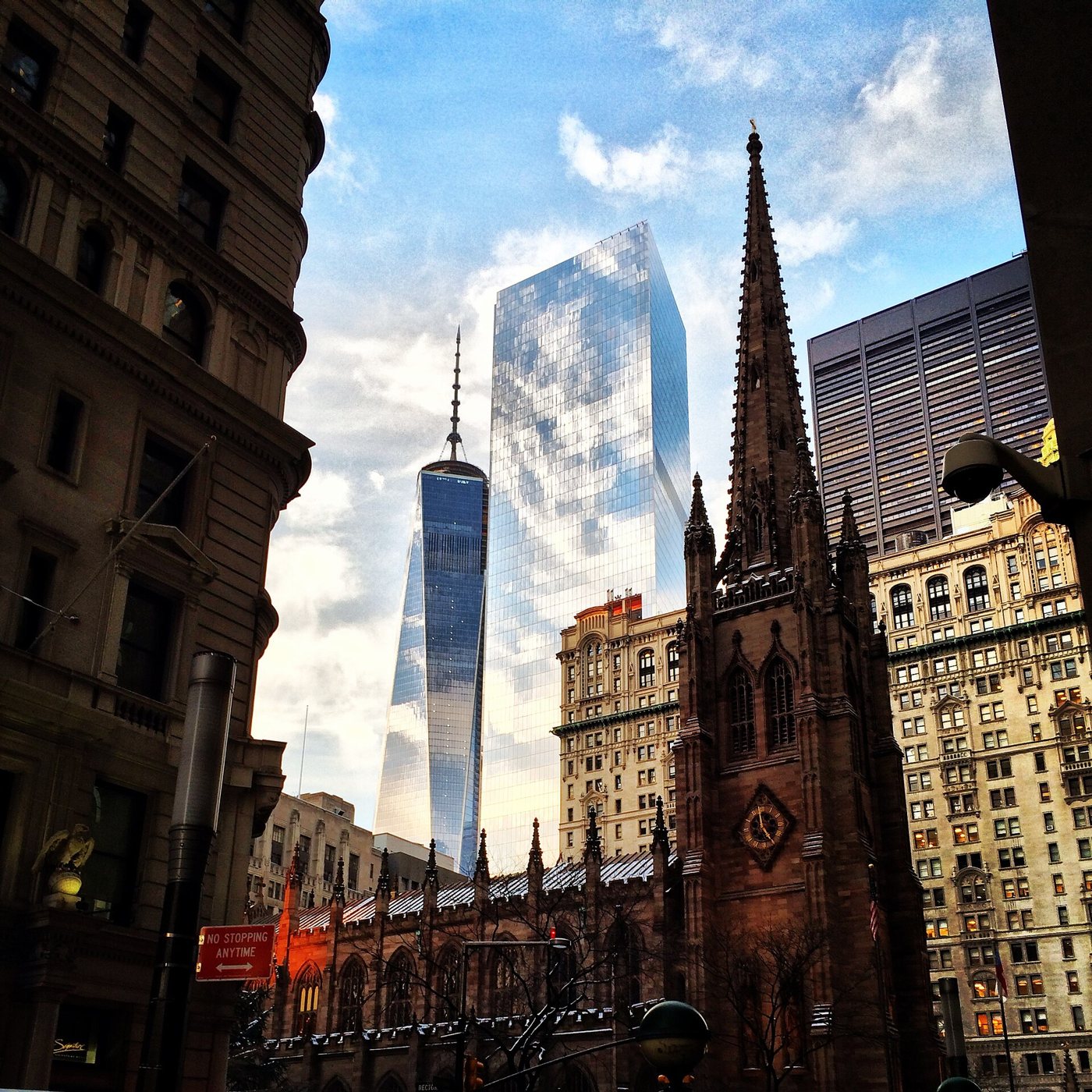 Trinity Church's brownstone spire and clock tower at Broadway and Wall Street, with the glass towers of the new World Trade Center rising behind it against a blue sky