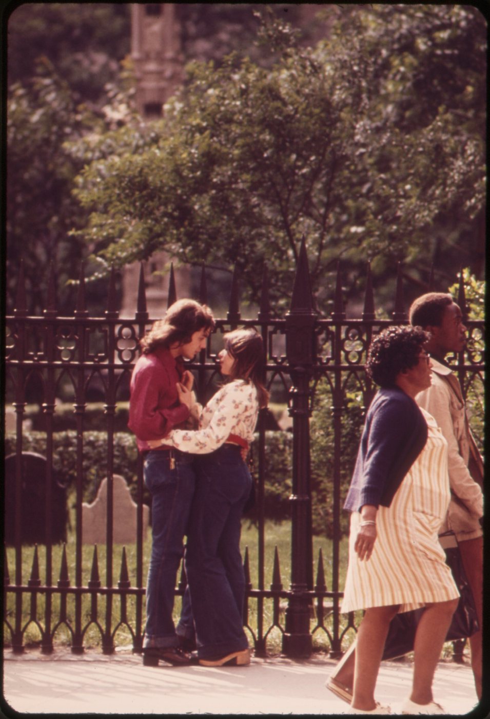 Two people embracing beside the black iron fence of Trinity Church's churchyard, old gravestones and trees behind them, two passers-by walking past on the right